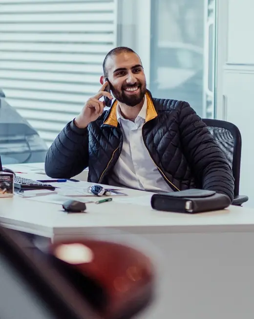 Lächelnder Autoverkäufer sitzt am Schreibtisch und telefoniert, Unterlagen und Autoschlüssel vor sich.