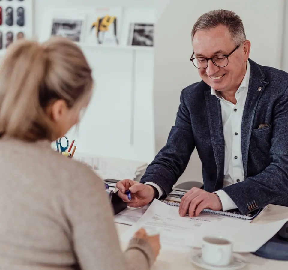 Mann im Anzug berät Kundin an einem Schreibtisch, beide blicken auf Vertragsunterlagen; im Vordergrund eine Kaffeetasse.