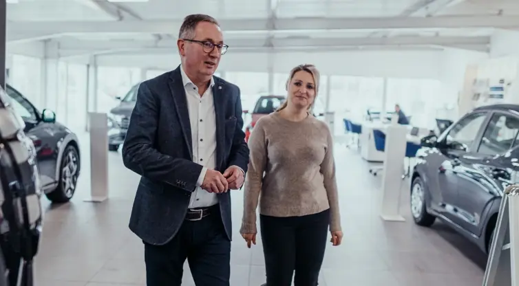 Mann im Anzug und Frau im Pullover gehen gemeinsam durch ein Autohaus zwischen ausgestellten Fahrzeugen