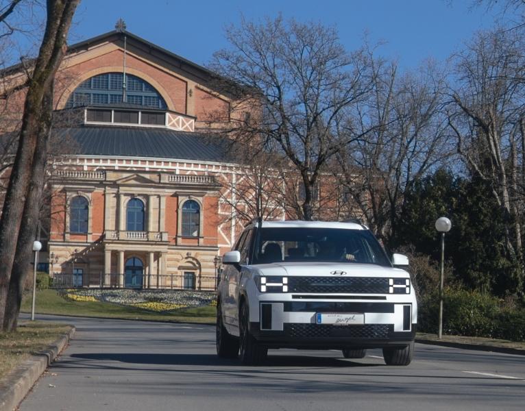 Weißes Hyundai Elektro-SUV fährt die Zufahrt vor dem Bayreuther Festspielhaus entlang, frontal aufgenommen bei sonnigem Wetter.