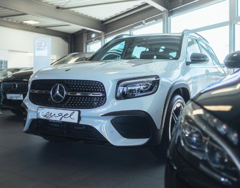 Weißer Mercedes-Benz SUV im Autohaus, Frontansicht mit Sterngrill und LED-Scheinwerfern; weitere Fahrzeuge im Hintergrund.