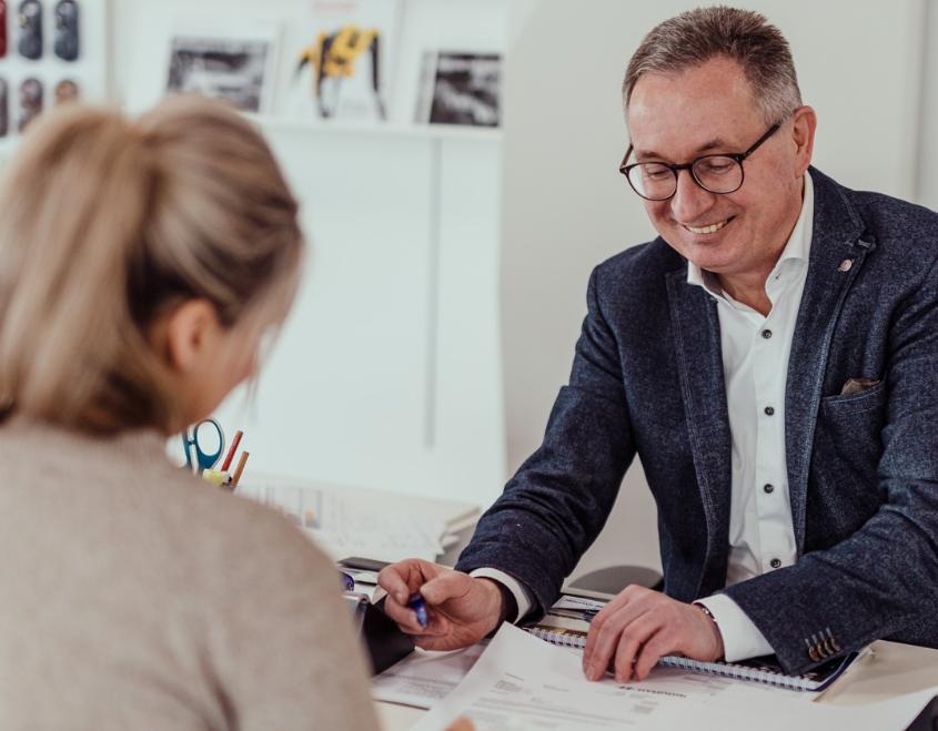 Autoverkäufer im Gespräch mit einer Kundin, Unterlagen und Verträge liegen auf dem Tisch