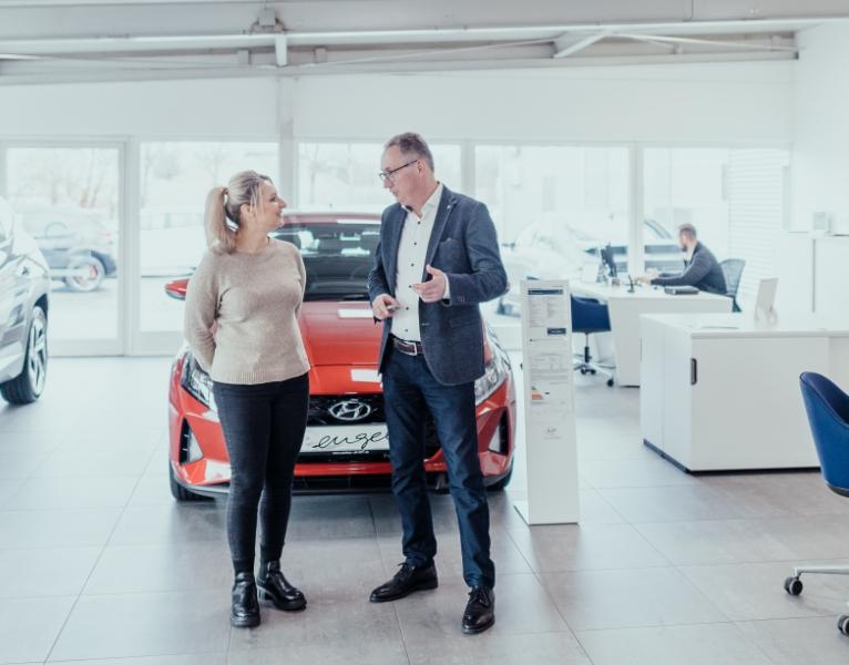 Kundin im Gespräch mit einem Autoverkäufer im Hyundai-Showroom, rotes Fahrzeug im Hintergrund