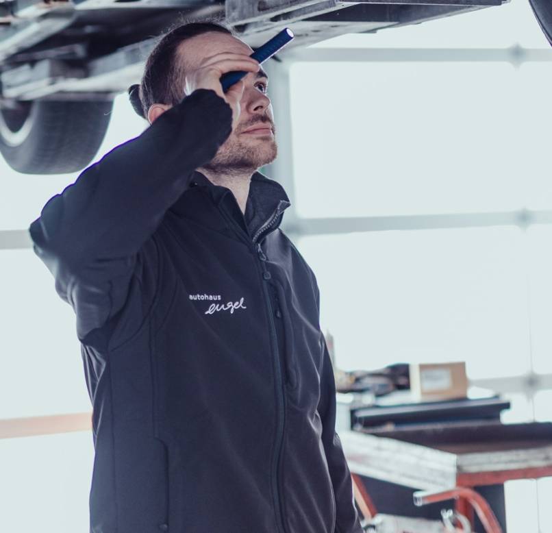Mechaniker in schwarzer Autohaus-Engel-Jacke prüft mit einer Taschenlampe den Unterboden eines angehobenen Fahrzeugs.
