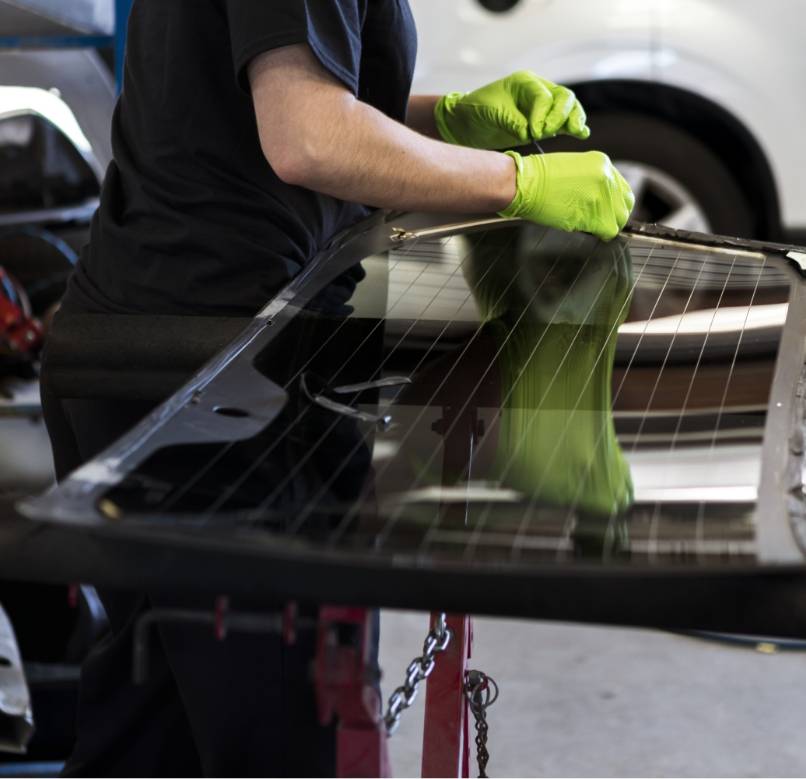 Mechaniker mit grünen Schutzhandschuhen arbeitet an einer ausgebauten Heckscheibe in der Werkstatt.