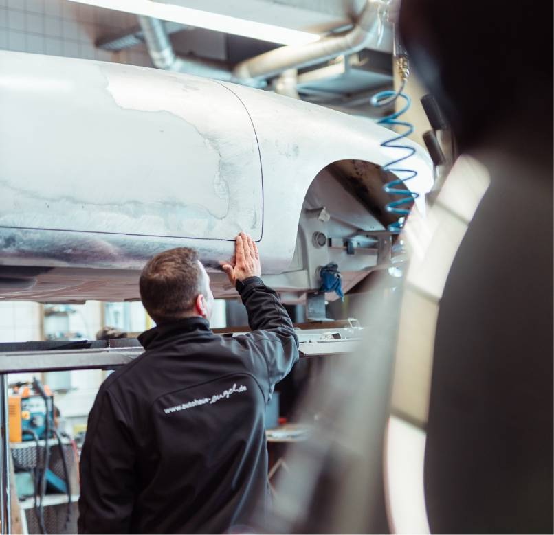 Mechaniker in schwarzer Autohaus-Engel-Jacke prüft mit der Hand die Karosserie eines aufgebockten Fahrzeugs in der Werkstatt.
