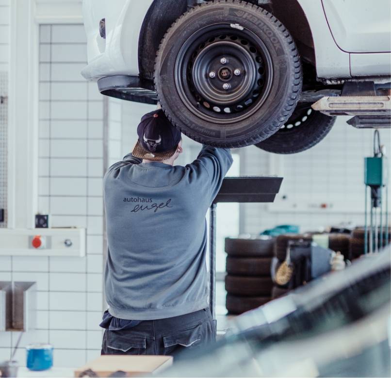 Mechaniker in grauem Autohaus-Engel-Pullover arbeitet an einem angehobenen Fahrzeugrad auf der Hebebühne in der Werkstatt.