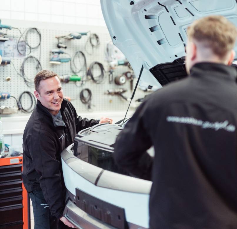 Zwei Mechaniker in Autohaus-Engel-Arbeitskleidung montieren gemeinsam eine Motorhaube in der Werkstatt.