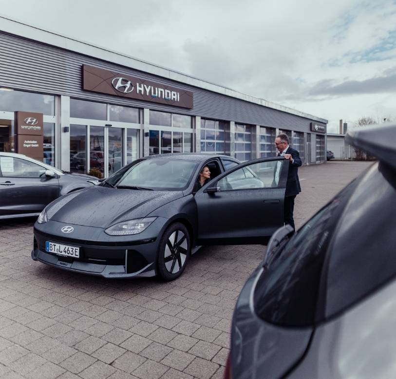 Mattschwarzes Hyundai Elektroauto vor dem Autohaus Engel; eine Frau sitzt am Steuer, ein Mitarbeiter hält die Tür geöffnet.
