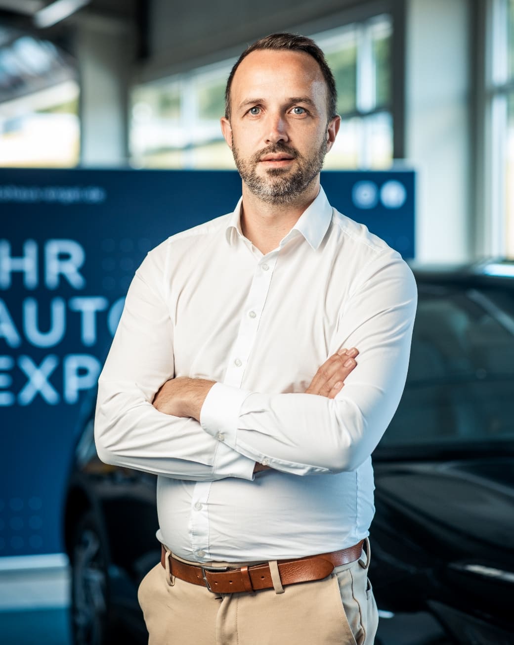 Ein Mann mit kurzem braunem Haar, Dreitagebart und weißem Hemd steht mit verschränkten Armen lächelnd in einem Autohaus. Im unscharfen Hintergrund sind ein Fahrzeug und ein blaues Schild mit der Aufschrift „Ihr Auto Experte“ zu sehen.